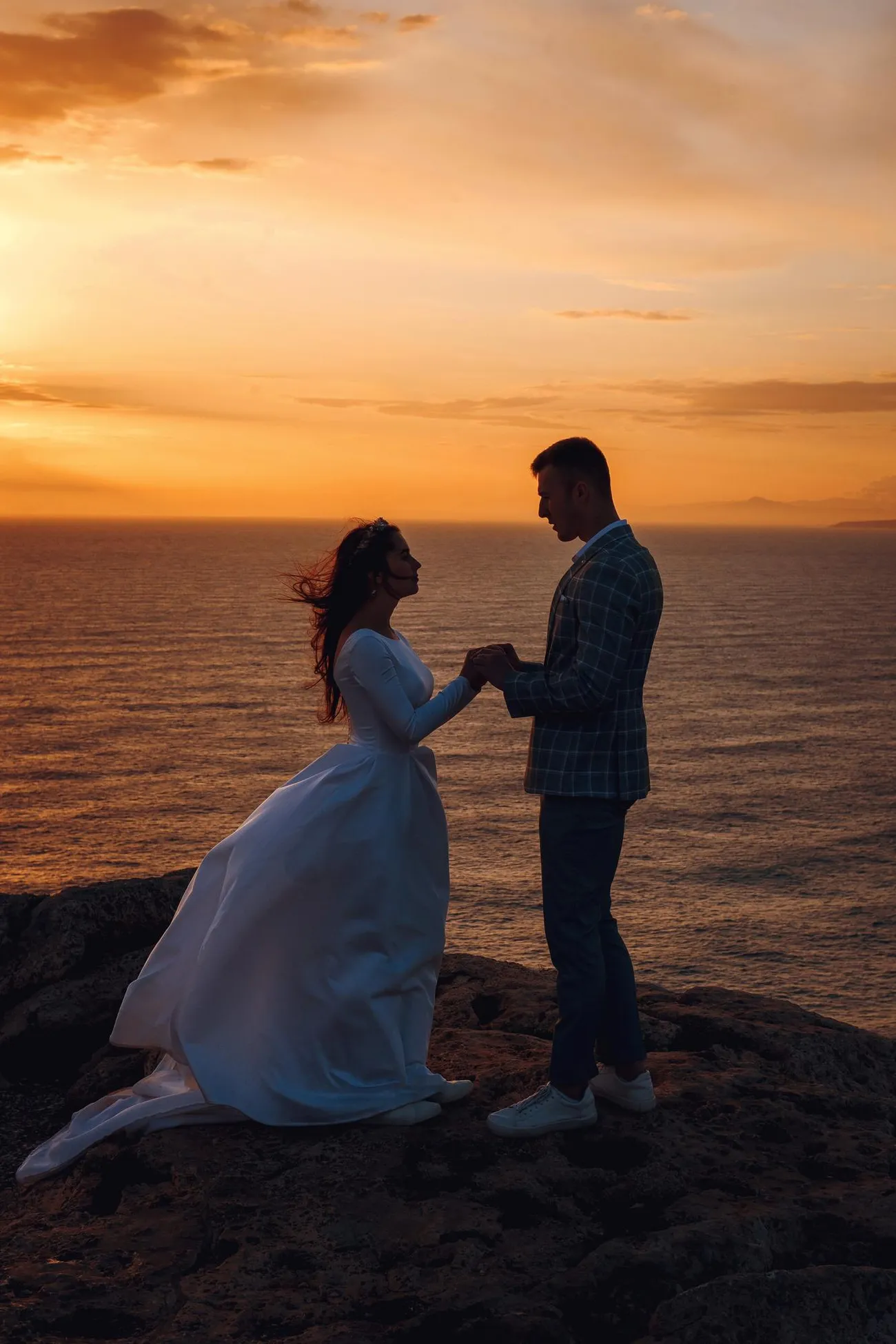 A couple in wedding attire holding hands on a coastal cliff at sunset